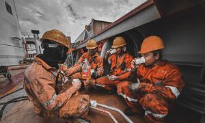 Four seafarers in orange coveralls and hard hats taking a break on the deck of a ship, drinking from cups and sharing a bottle of drink.
