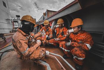 Four seafarers in orange coveralls and hard hats taking a break on the deck of a ship, drinking from cups and sharing a bottle of drink.