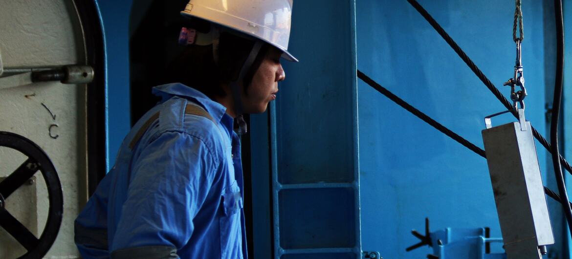 A seafarer wearing blue coveralls and a white safety helmet standing in a ship's engine room.