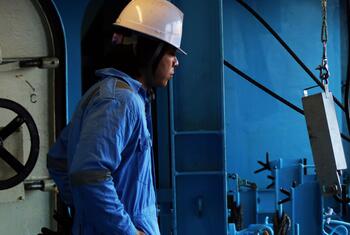 A seafarer wearing blue coveralls and a white safety helmet standing in a ship's engine room.