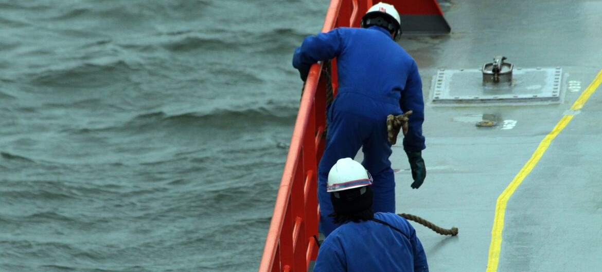 Two seafarers in blue jumpsuits and white hard hats on the deck of a ship, near the railing with the ocean in the background.
