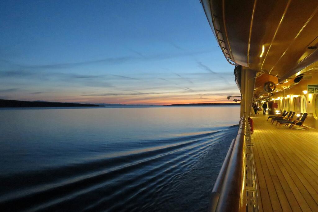 Vista de un pasajero desde la cubierta de un crucero al atardecer, mostrando la vigilia en el mar tranquilo mientras viaja hacia la lejana costa de Alaska bajo un colorido cielo crepuscular.