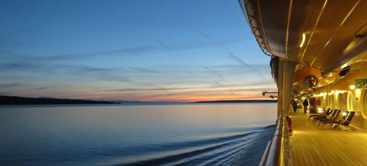 A passenger's view from the deck of a cruise ship at dusk, showing the wake in the calm sea as it travels towards the distant Alaskan coastline under a colorful twilight sky.