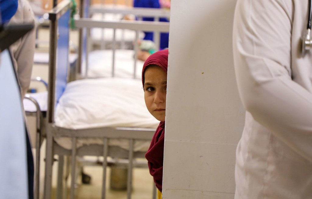 Msichana wa Afghanistan aliyevaa kitambaa chekundu kichwani anachungulia kutoka nyuma ya ukuta katika wodi ya hospitali, huku vitanda vya hospitali na wafanyakazi wakionekana nyuma.