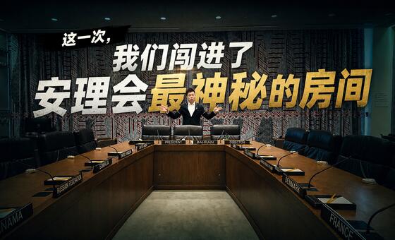 一名身着西装的男子站在联合国安理会会议室内的一张大型会议桌旁，桌上可见各国名称牌。