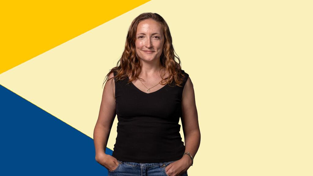 Una mujer con cabello largo, ondulado y de color castaño rojizo, vestida con una blusa negra sin mangas y jeans, posa sonriendo frente a un fondo geométrico diagonal amarillo y azul.