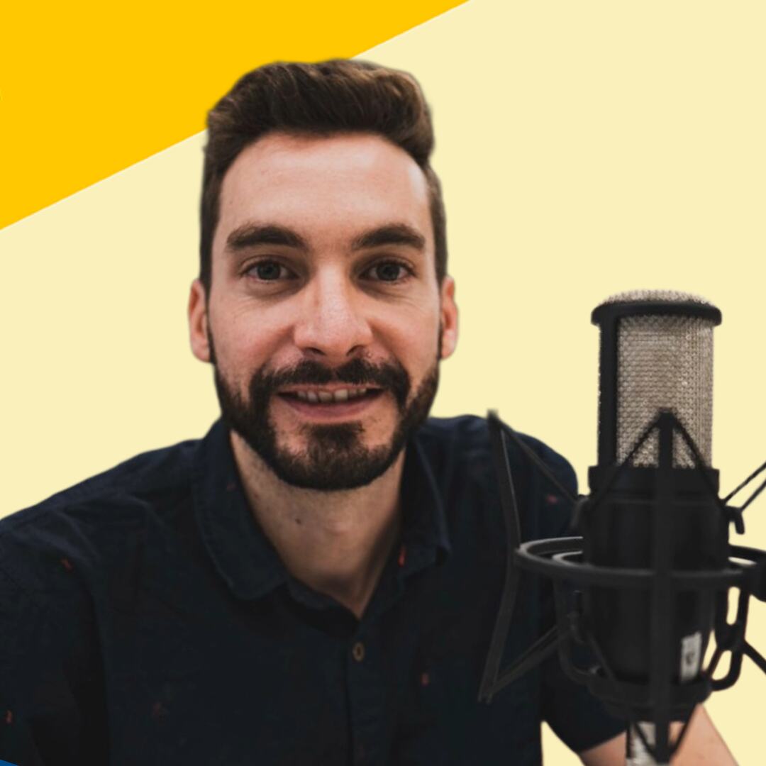 Un hombre barbudo sonriente, con una camisa azul marino oscura, está sentado frente a un micrófono de condensador de estudio, sobre un fondo diagonal amarillo y crema.