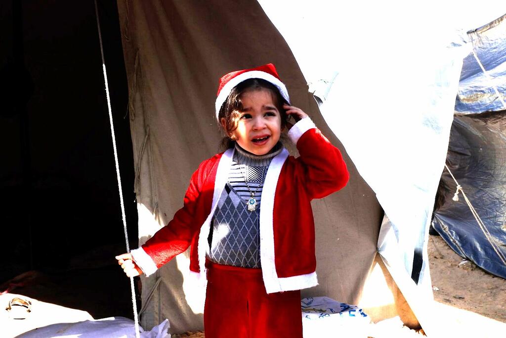 A young Palestinian girl wearing a Santa Claus outfit, standing in front of tents of displaced families in northern Gaza City.