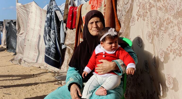 Na‘ma Al-Khawaja, an elderly Palestinian woman, carrying her granddaughter who is wearing a Christmas outfit, in front of their tent in northern Gaza City.