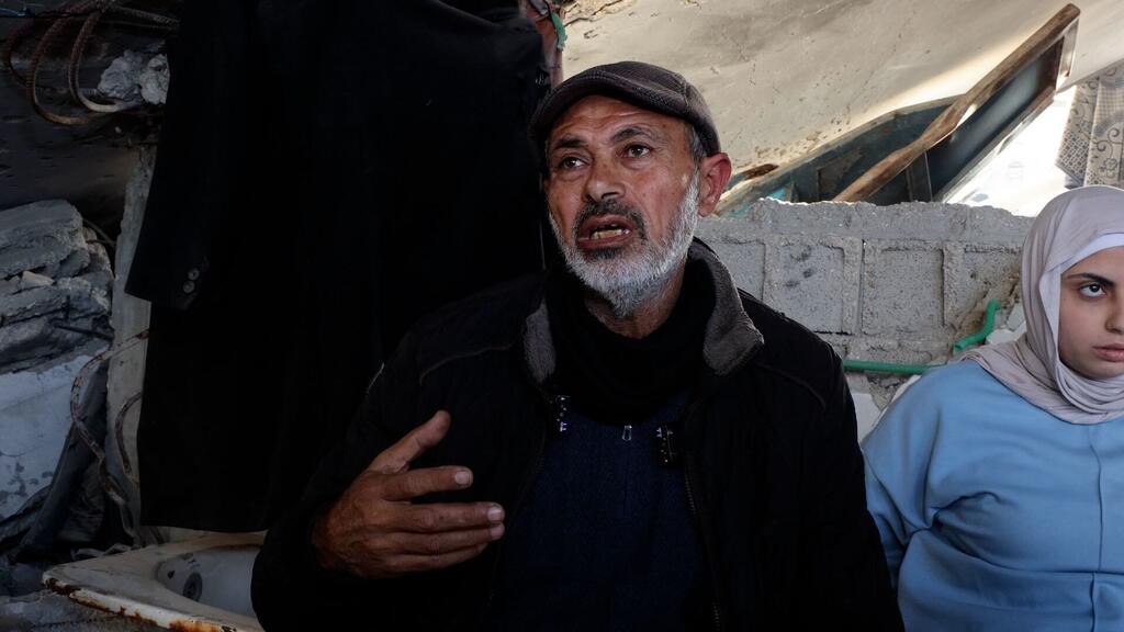 Un hombre de Oriente Medio con barba gris habla mientras hace gestos, de pie junto a una joven palestina con hiyab en medio de escombros y estructuras dañadas, destacando el impacto en la comunidad.