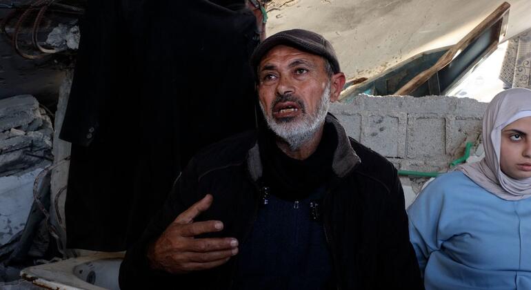 رجل من الشرق الأوسط ذو لحية رمادية يتحدث مع إشارات، يقف بجوار امرأة فلسطينية شابة في حجاب وسط الأنقاض والهياكل المتضررة، ويبرز التأثير على المجتمع.