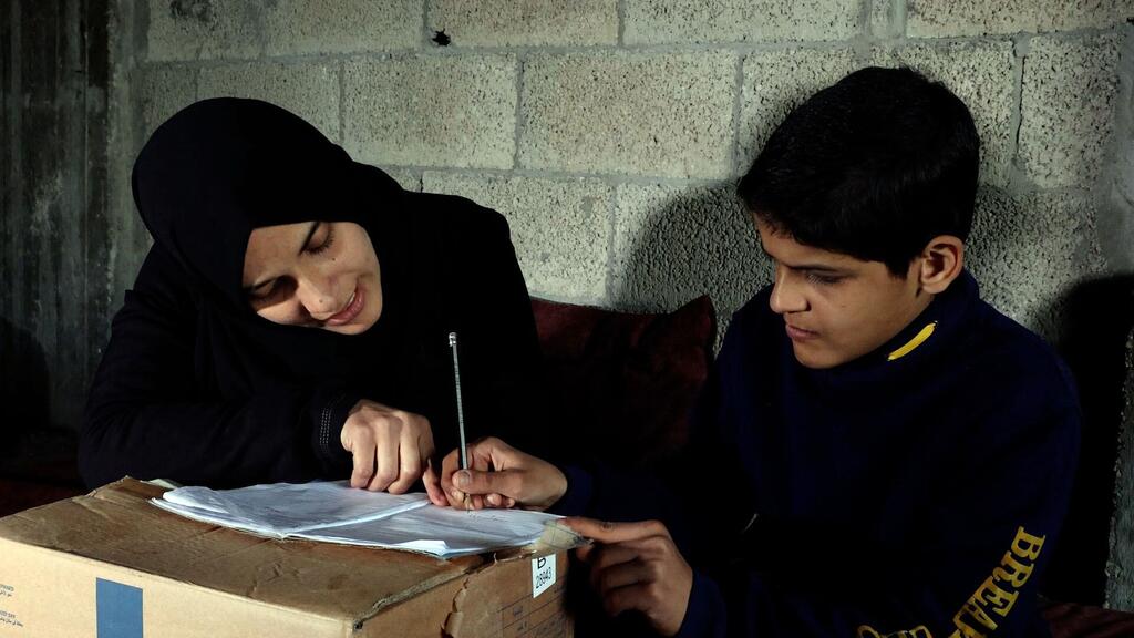 Una mujer palestina que lleva un hijab negro ayuda a un niño pequeño con sus deberes escolares, escribiendo en un pedazo de papel que descansa sobre una caja de cartón.