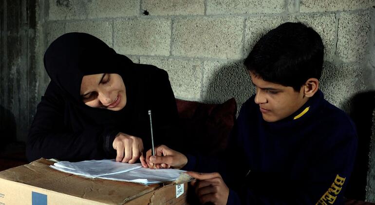امرأة فلسطينية ترتدي حجابًا أسود تبتسم بينما تساعد صبيًا صغيرًا في واجباته المدرسية، وتكتب على قطعة ورق تقع على علبة ورقية.