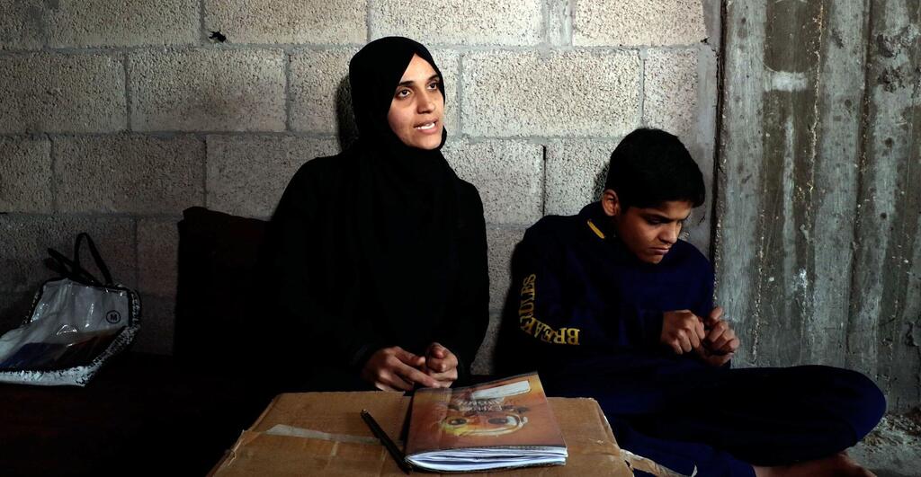 Una mujer palestina y un niño con autismo se sientan en un escritorio improvisado en una habitación sencilla con paredes de bloques de cemento, enfocados en materiales de aprendizaje.