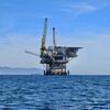 Офшорная нефтяная платформа стоит в голубом океане под чистым небом, с горами, видимыми на расстоянии.