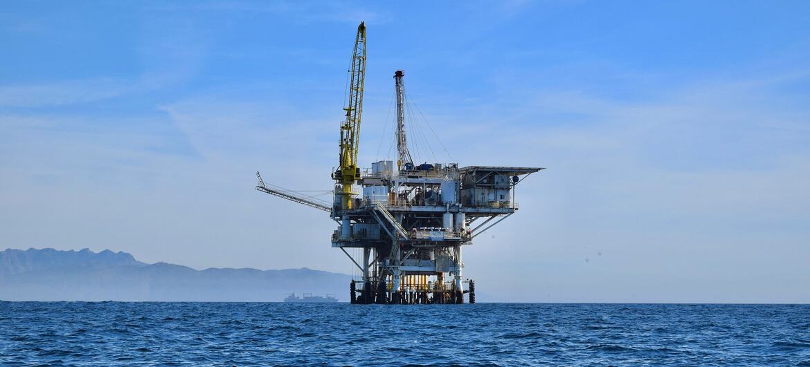 Une plate-forme pétrolière offshore se dresse dans l'océan bleu sous un ciel clair, avec des montagnes visibles au loin.