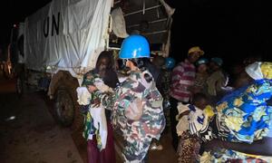 A female UN peacekeeper in camouflage uniform and blue helmet assists a woman holding a child near a UN truck at night in Ituri, DRC.