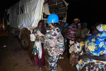 Une femme de l'ONU en uniforme de camouflage et casque bleu aide une femme tenant un enfant près d'un camion de l'ONU la nuit à Ituri, en RDC.