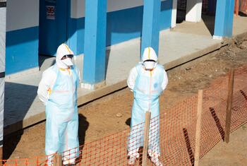 Two healthcare workers in protective gear stand outside a blue building under construction, enclosed by an orange safety fence in Bunia, DR Congo.