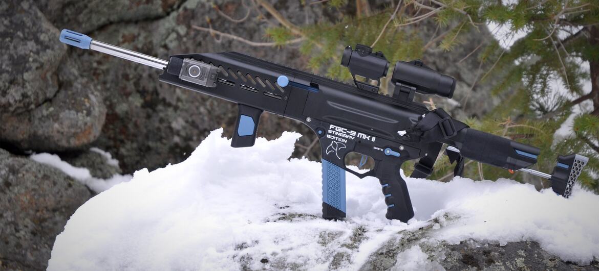 A black and blue FGC-9 MKII Stingray airsoft rifle rests on a snow-covered rock with pine branches in the background.