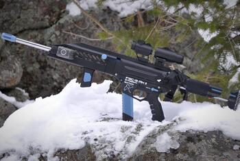 Un rifle de airsoft FGC-9 MKII Stingray negro y azul descansa sobre una roca cubierta de nieve con ramas de pino al fondo.
