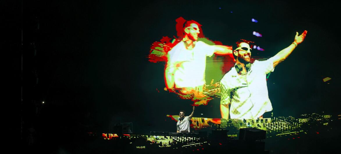 Um DJ se apresenta em um palco escuro com uma grande tela atr&aacute;s dele exibindo uma imagem estilizada e art&iacute;stica de dois artistas.