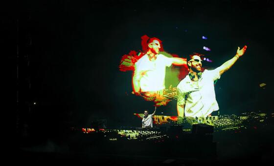 Um DJ se apresenta em um palco escuro com uma grande tela atrás dele exibindo uma imagem estilizada e artística de dois artistas.