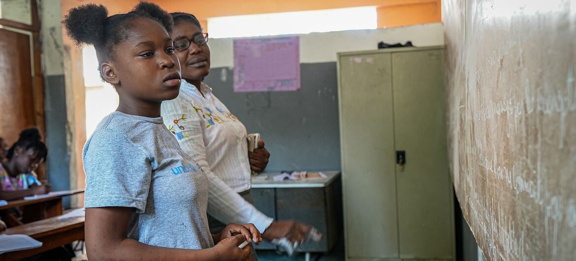 Dieussika, a displaced child in Haiti, stands in her classroom wearing a UNICEF shirt, holding a chalk, and appears focused as she participates in a lesson. She credits UNICEF's catch-up classes and vocational training for helping her return to school and graduate, emphasizing the importance of not abandoning children.