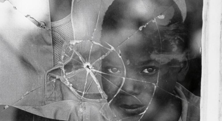 Uma criança refugiada olha através de uma janela quebrada, com o rosto marcado por vidro quebrado, refletindo as duras realidades do deslocamento em Moçambique.