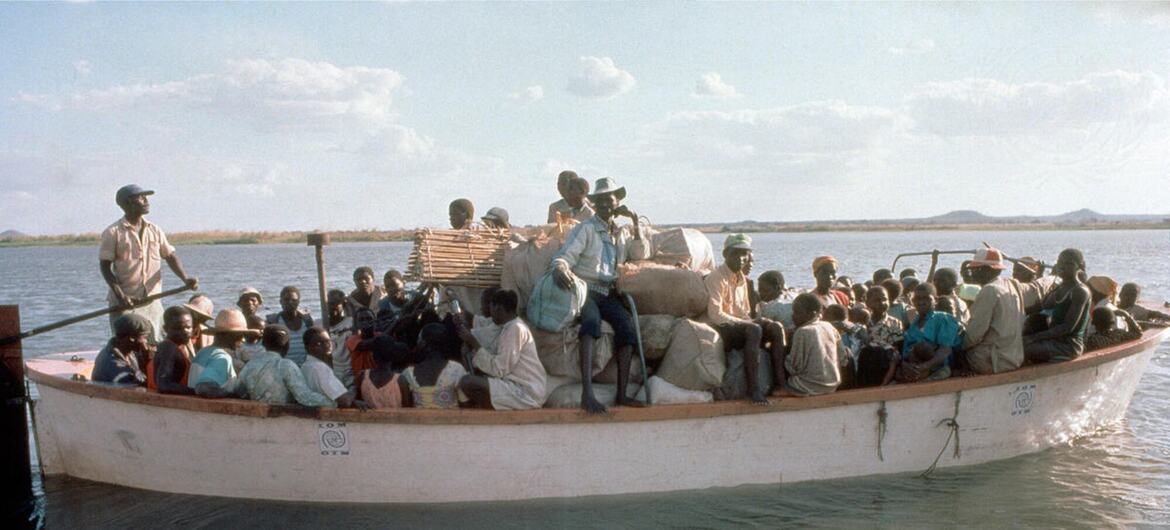 Um barco lotado transportando refugiados e suas pertences na água, com um homem dirigindo na frente.