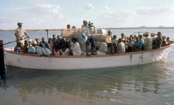 Um barco lotado transportando refugiados e suas pertences na água, com um homem dirigindo na frente.