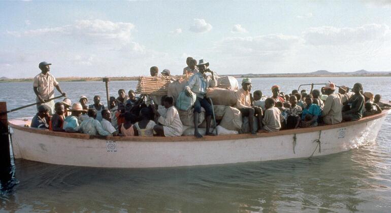 Um barco lotado transportando refugiados e suas pertences na água, com um homem dirigindo na frente.