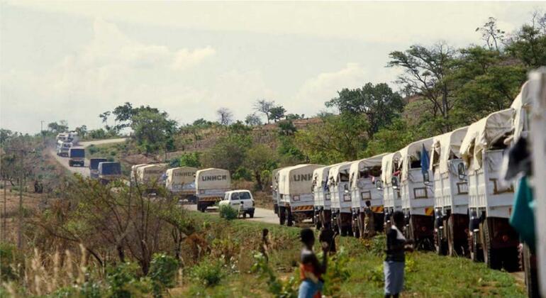 Uma linha de caminhões de ajuda do ACNUR percorre uma estrada rural em Moçambique, com crianças de pé perto em uma área ereta.