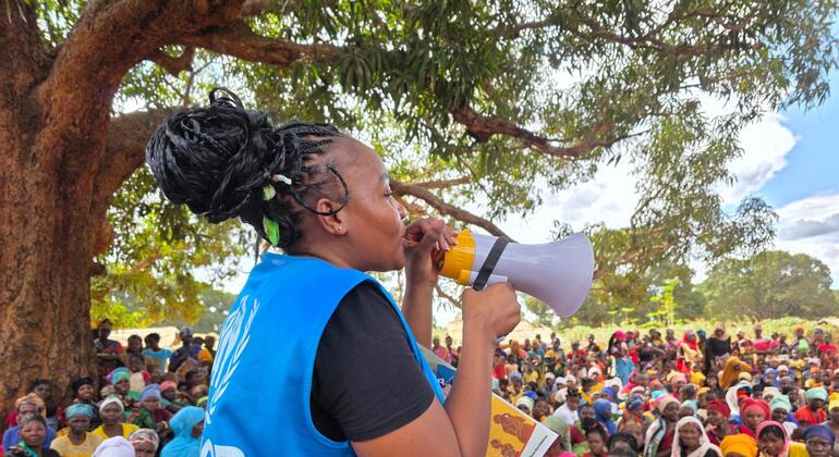 Uma trabalhadora humanitária do ACNUR usa um megafone para dirigir-se a uma grande multidão de pessoas deslocadas em um ambiente exterior sombreado por árvores em Moçambique.