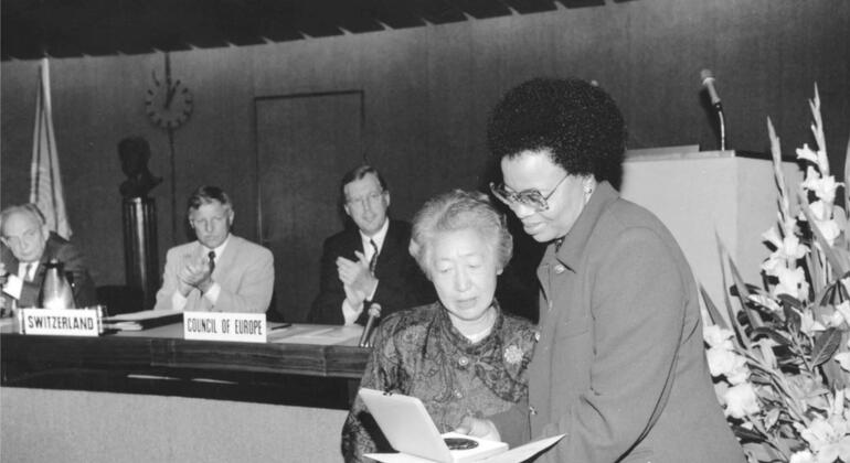 Uma mulher vestida de fato entrega um prémio a uma mulher idosa num evento formal, com diplomatas da Suíça e do Conselho da Europa aplaudindo no fundo.
