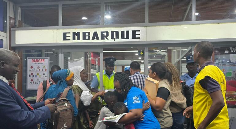 Um grupo de refugiados, incluindo mulheres e crianças, está reunido numa área de embarque do aeroporto marcada "EMBARQUE", com o apoio do pessoal do ACNUR.