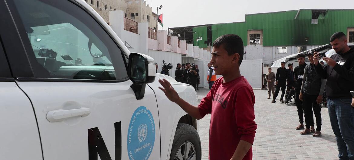 A young boy in a red shirt gestures toward a white UN humanitarian aid vehicle parked outside Al-Amal Hospital in Gaza, with people and buildings in the background.
