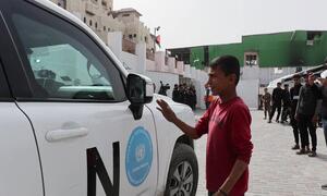 A young boy in a red shirt gestures toward a white UN humanitarian aid vehicle parked outside Al-Amal Hospital in Gaza, with people and buildings in the background.