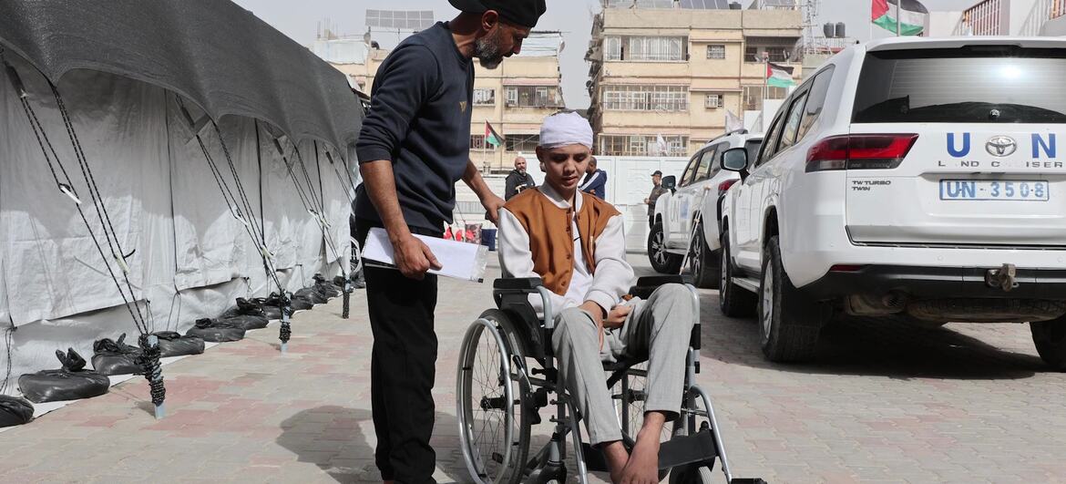 A patient in a wheelchair is being assisted by a staff member at Al-Amal Hospital in Gaza, affiliated with the Palestinian Red Crescent Society, preparing for transport. A UN vehicle is parked nearby.
