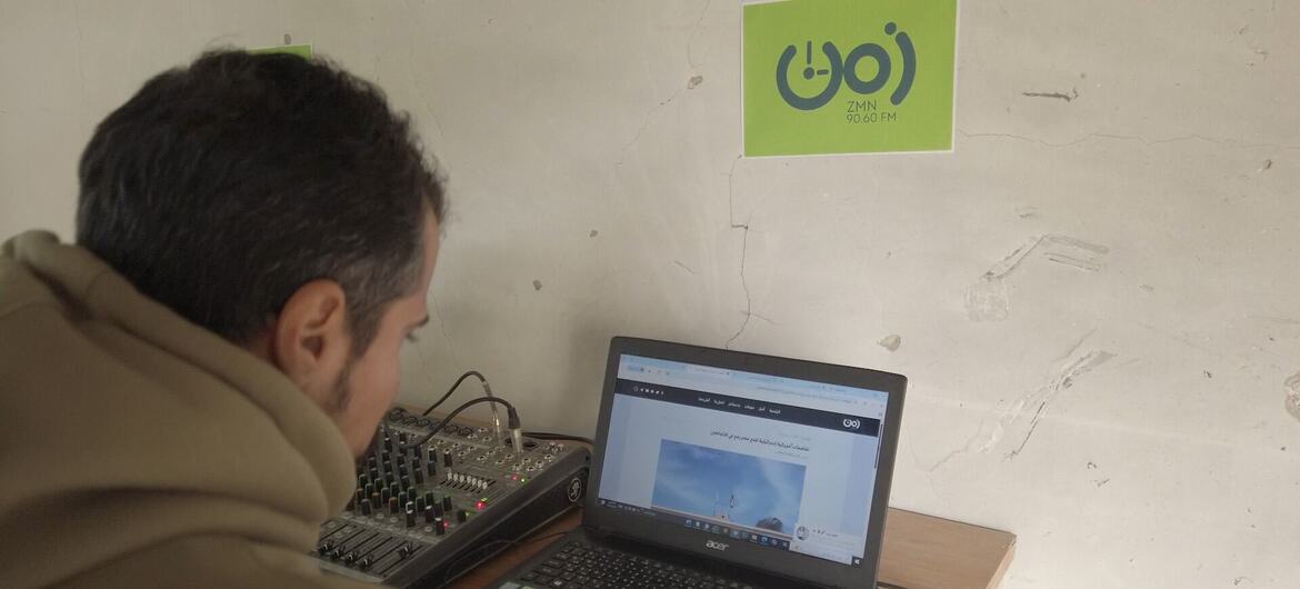 Un hombre está transmitiendo desde una estación de radio en Gaza, usando una computadora portátil y un mezclador de audio con un logotipo ZMN 90.60 FM en la pared.