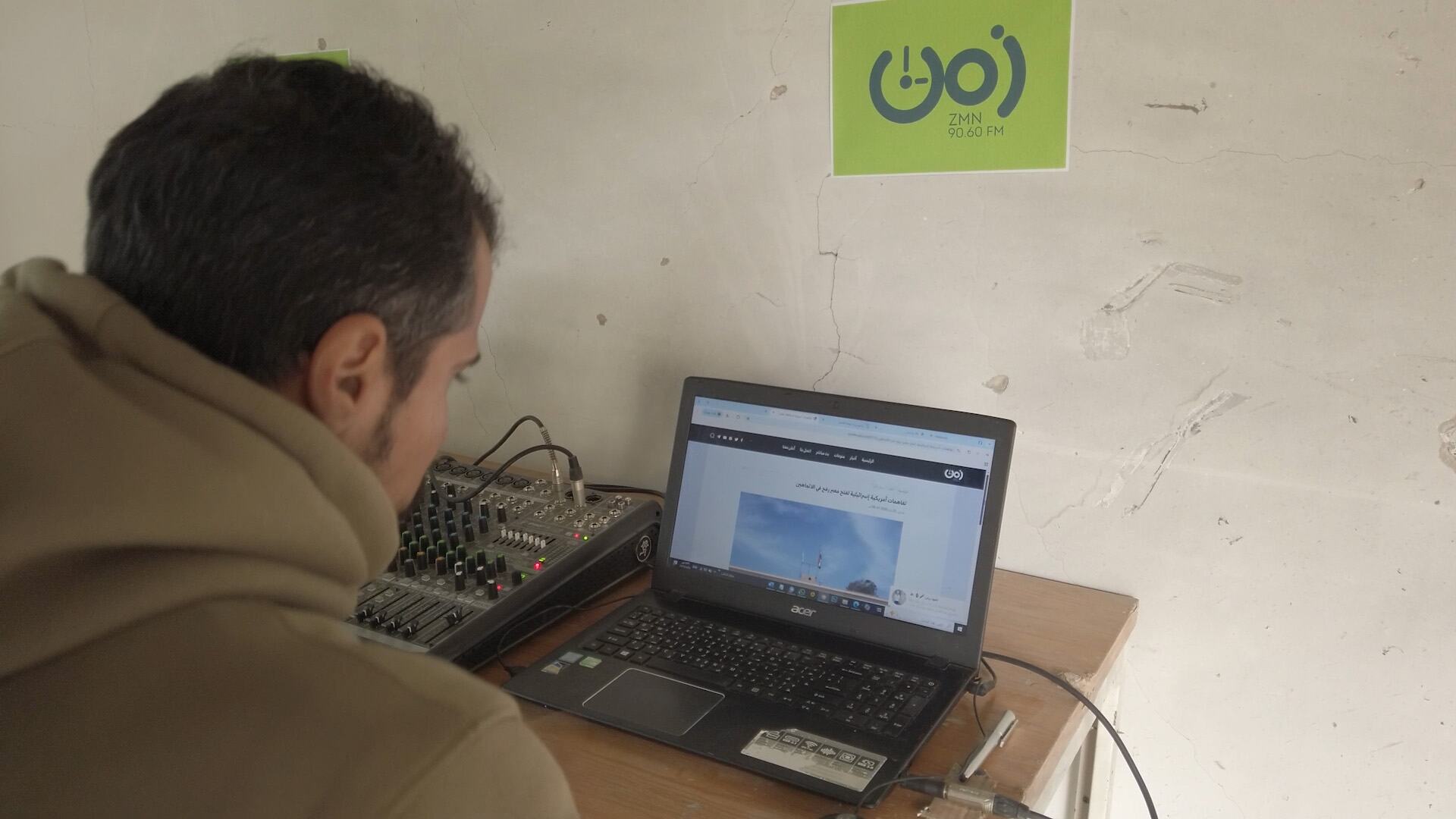 Un homme diffuse depuis une station de radio à Gaza, à l'aide d'un ordinateur portable et d'un mélangeur audio avec un logo ZMN 90.60 FM sur le mur.