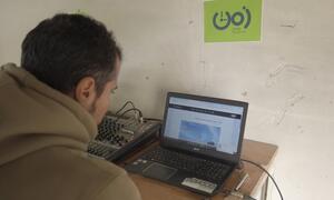 Un hombre está transmitiendo desde una estación de radio en Gaza, usando una computadora portátil y un mezclador de audio con un logotipo ZMN 90.60 FM en la pared.