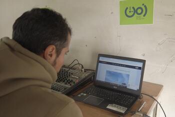 Un hombre está transmitiendo desde una estación de radio en Gaza, usando una computadora portátil y un mezclador de audio con un logotipo ZMN 90.60 FM en la pared.
