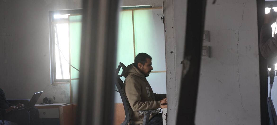 A journalist works at a desk in a damaged office in Gaza, viewed through broken pillars. Another person uses a laptop in the background.