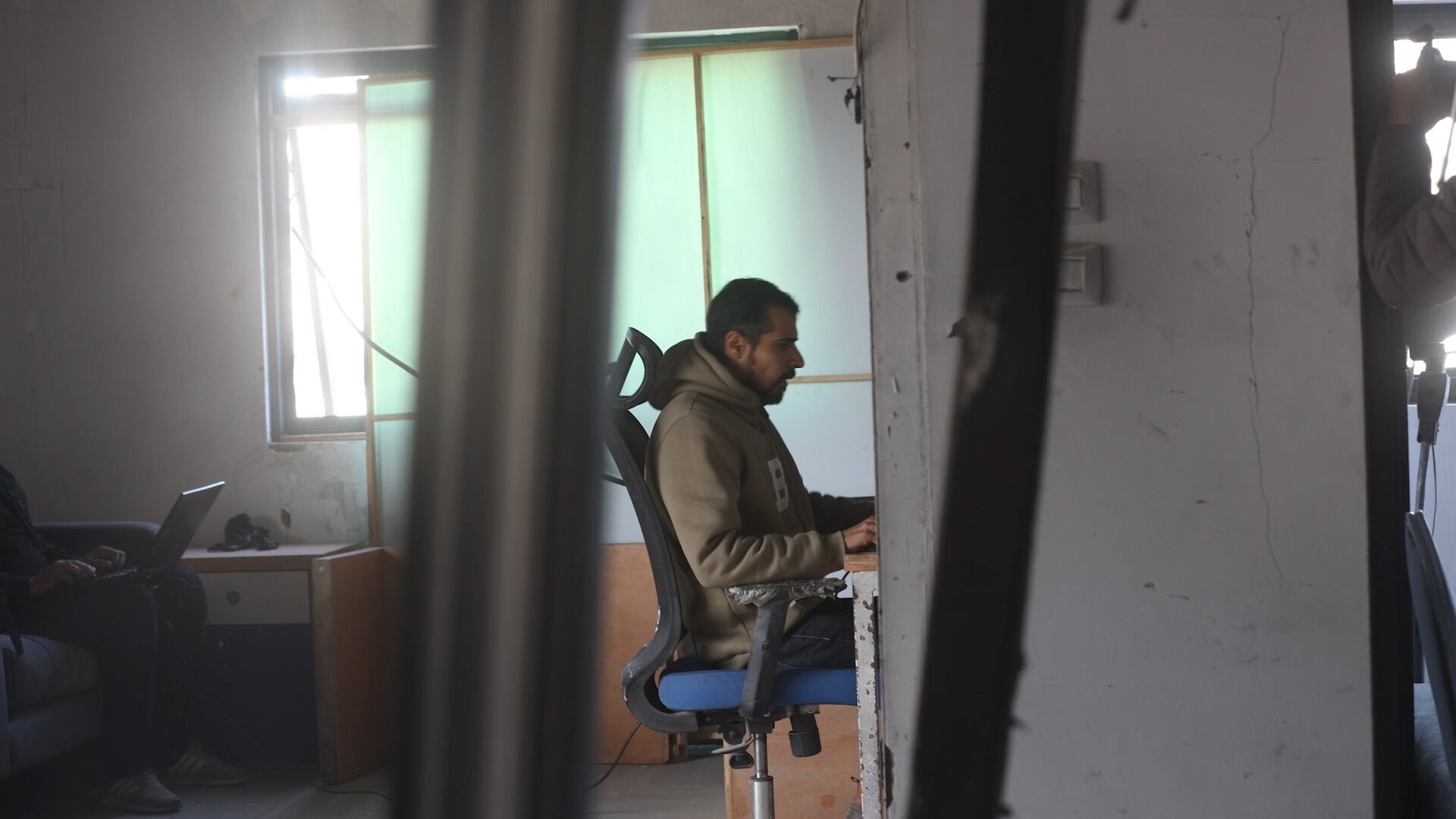 Un journaliste travaille à son bureau dans un bureau endommagé à Gaza, vu à travers des piliers brisés, une autre personne utilise un ordinateur portable en arrière-plan.