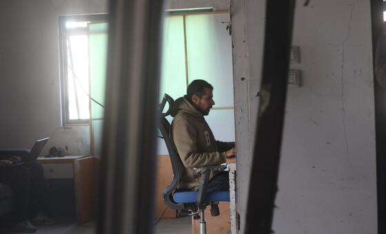 Un periodista trabaja en un escritorio en una oficina dañada en Gaza, visto a través de pilares rotos.