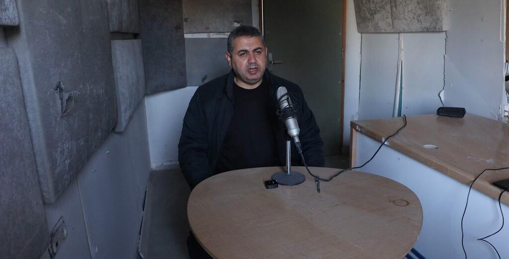 Un hombre está sentado en una mesa con un micrófono en una habitación dañada, probablemente llevando a cabo una entrevista en Gaza.