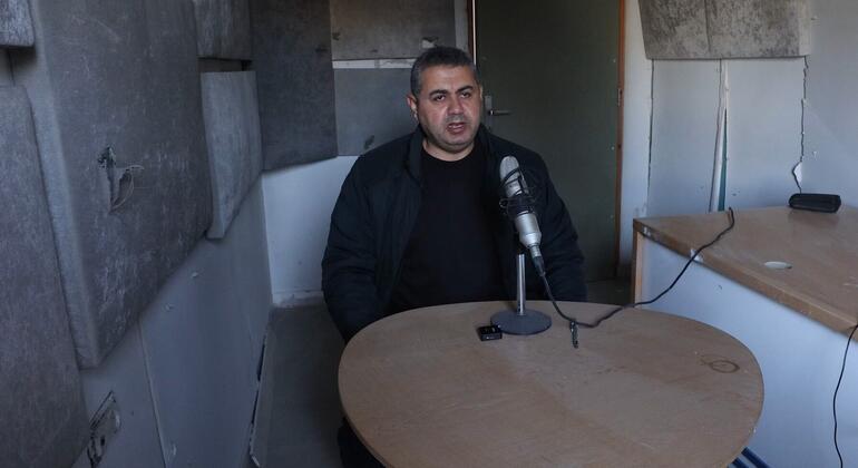 رجل يجلس على طاولة مع الميكروفون في غرفة مدمرة، وربما يجري مقابلة في غزة.