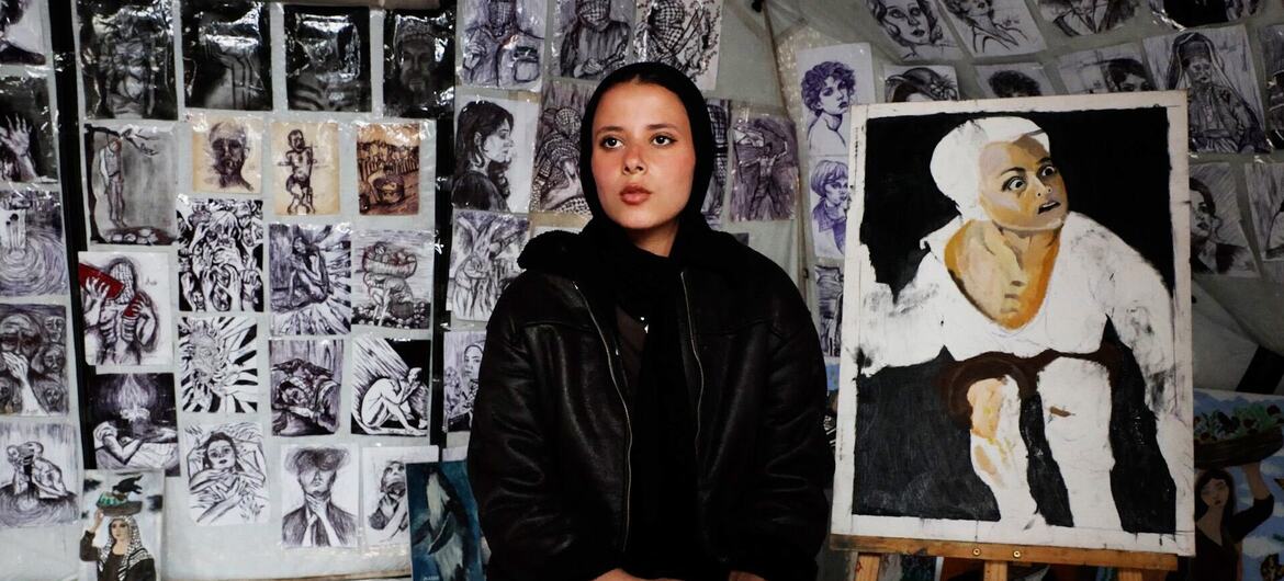 الفنانة الفلسطينية الشابة، فرح خالد تجلس أمام إحدى لوحاتها في الخيمة التي حولتها إلى معرض فني في قلب مدرسة تابعة لوكالة الأونروا التي تحولت إلى مركز لإيواء النازحين في مدينة غزة.