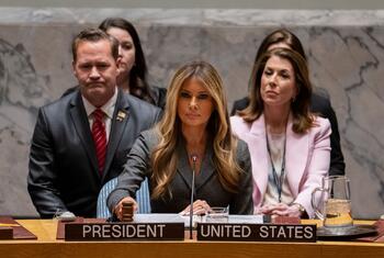 Melania Trump speaking at a United Nations meeting, seated at a desk with a 'President United States' placard.